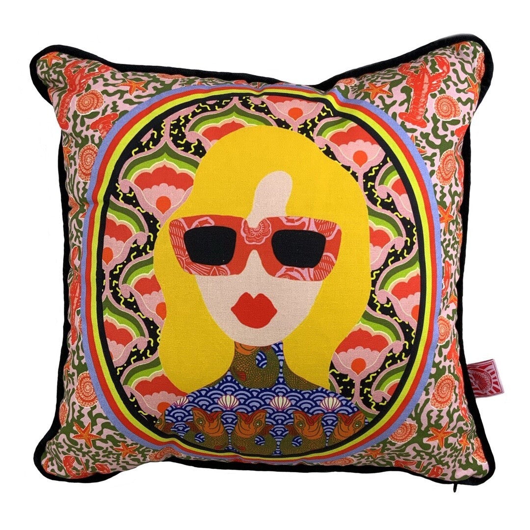 A cushion with an illustration of a seventies-inspired lady's face with red lips and sunglasses and blonde hair circled within a floral print with an illustrated lobster background.