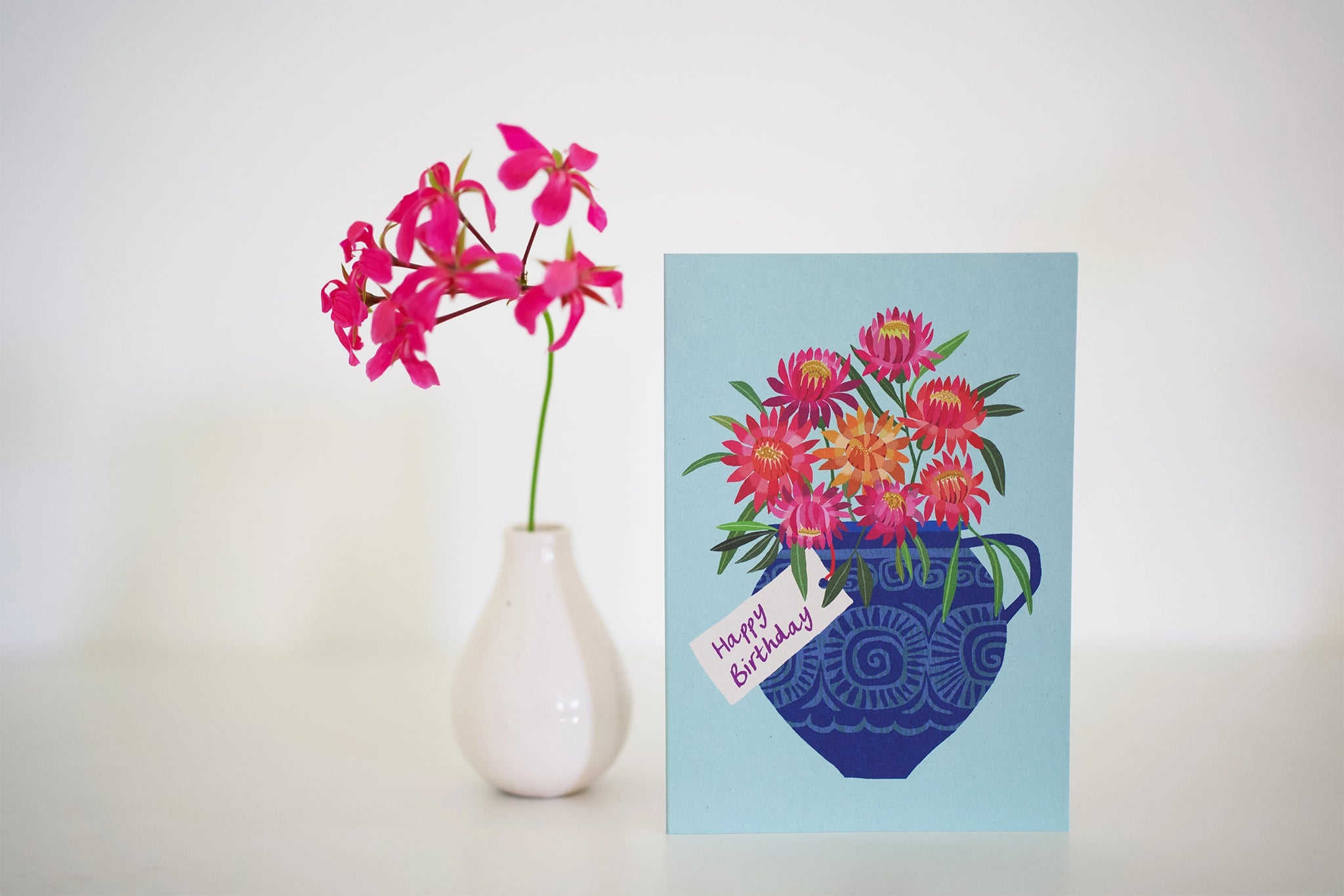 Vase with pink flowers next to an aqua card with floral design and 'Happy Birthday' text on a white background.