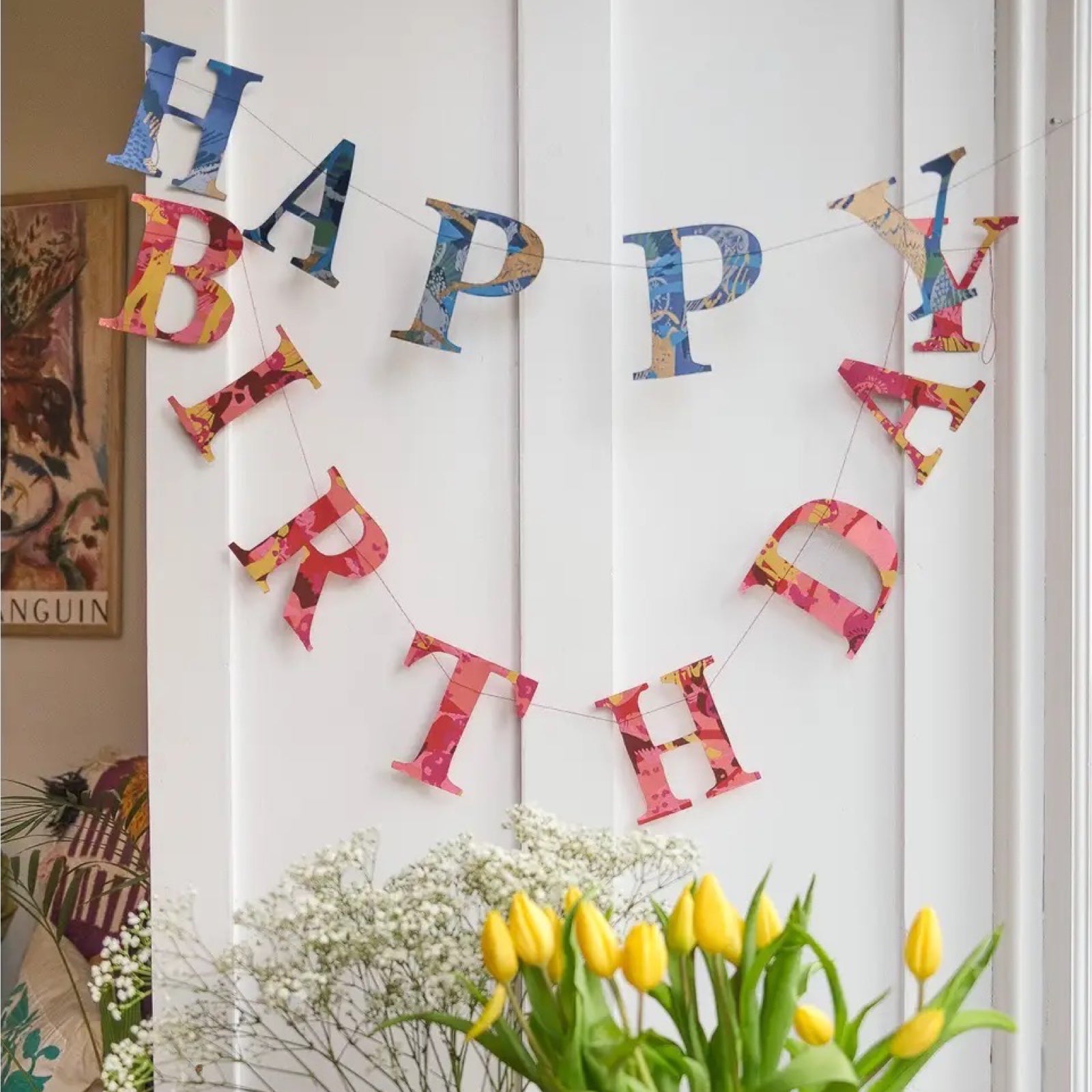 A happy birthday paper garland in red, and blue patterned letters hanging against a white wall.