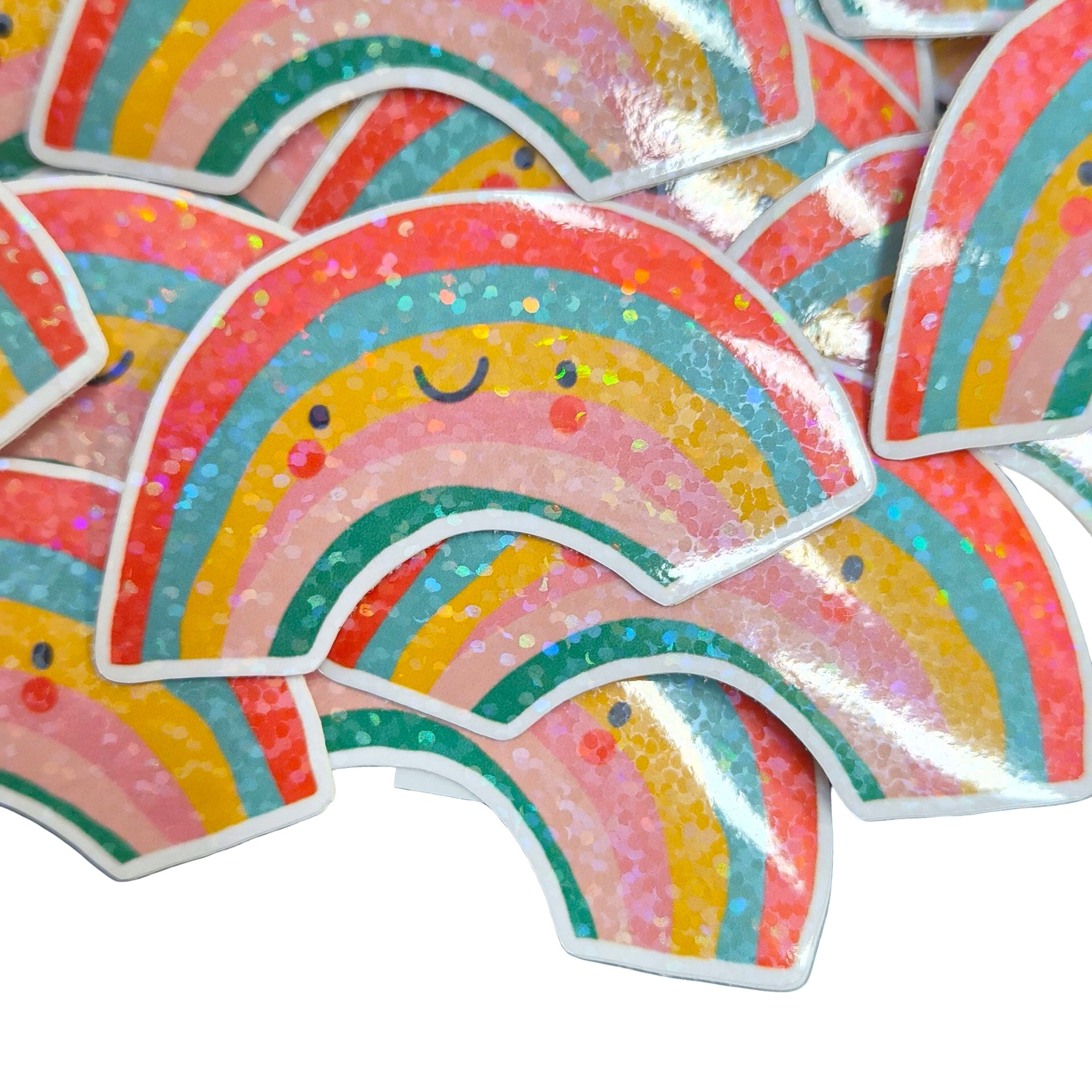 A pile of cute, smiling rainbow stickers with a glittery finish, featuring pastel stripes of red, orange, yellow, green, blue, and pink with white borders.