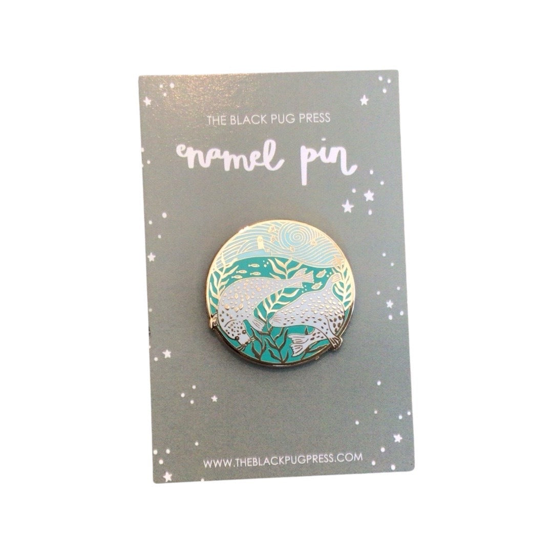 A circular enamel pin badge featuring an illustration of a couple of white seals swimming in a harbour.