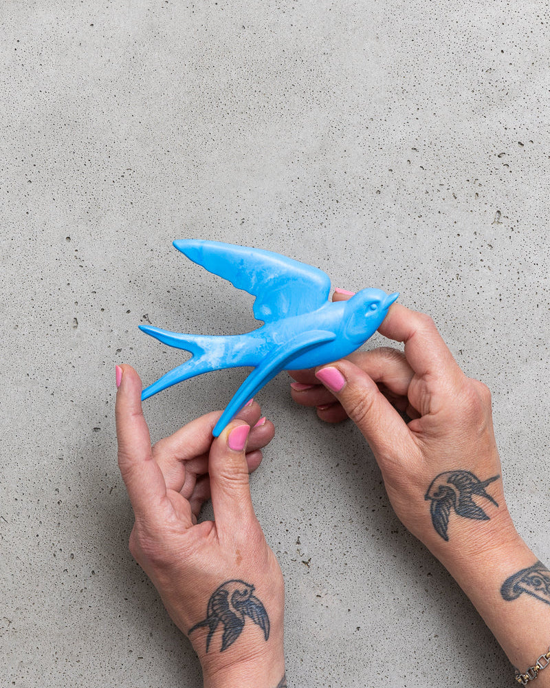 Blue bird-shaped object held by hands with tattoos on a grey surface.