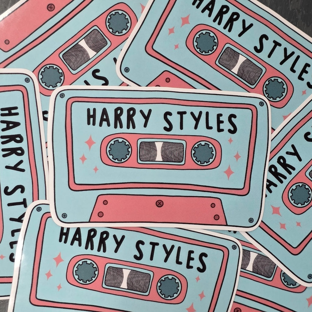 A rectangular-shaped sticker featuring an illustration of a retro cassette tape in light blue and pink with the title 'Harry Styles', decorated with pink stars, in a pile.