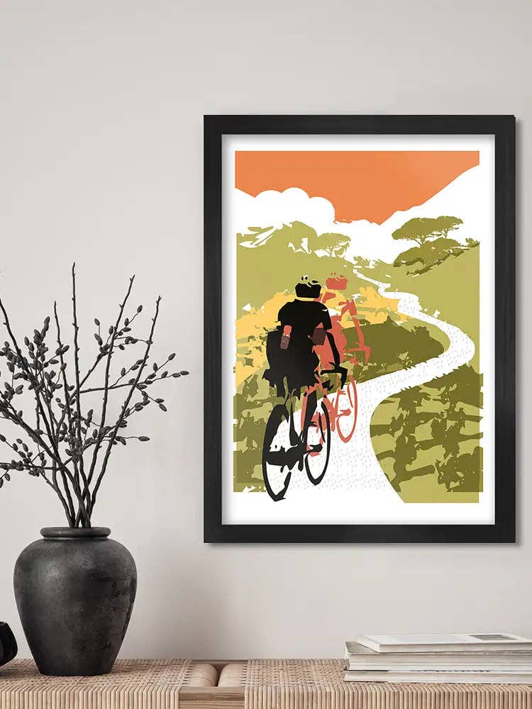 Framed artwork of a cyclist on a mountain path with a vase and books on a shelf below.