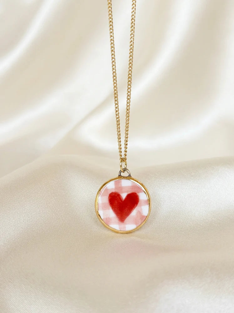 A gold chain necklace with a round pendant featuring a red heart on a pink and white checkered background, displayed on a draped cream satin fabric.