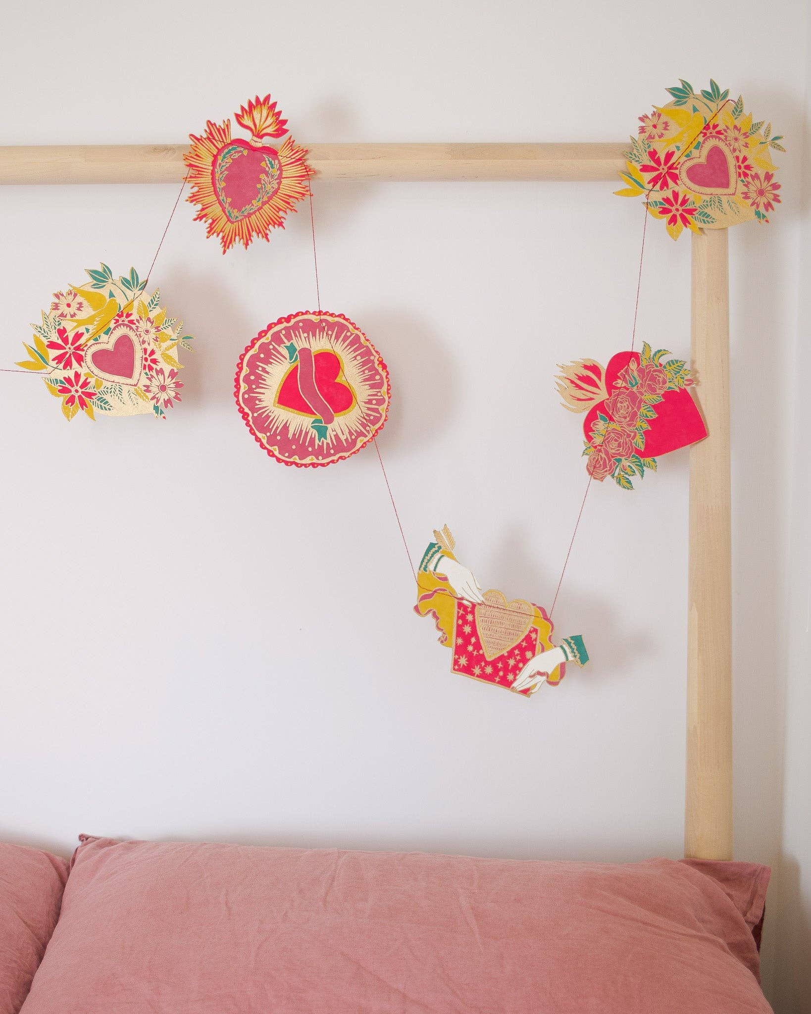 A screen printed paper garland with 10 individual heart designs in red, gold and green hanging along a wooden bed frame.