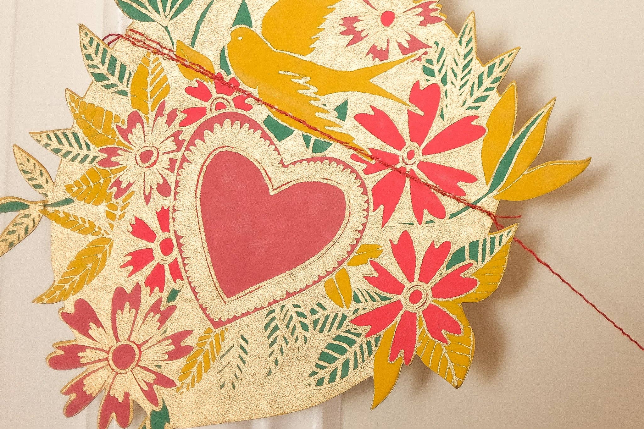 A heart design from a hearts paper garland with a red love heart in the centre surrounded by red flowers, green and mustard leaves, a mustard bird and gold details.