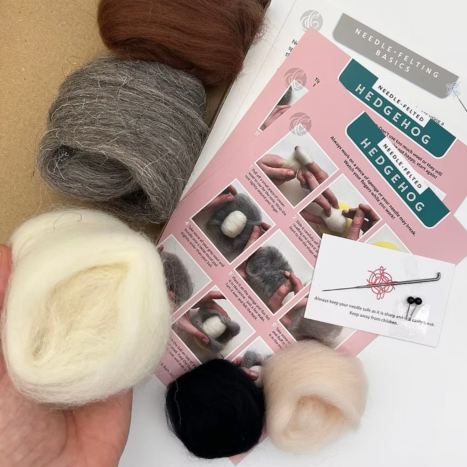 Ball of white yarn, brown yarn, grey yarn, and black yarn with a needle-felting guide on a white background.