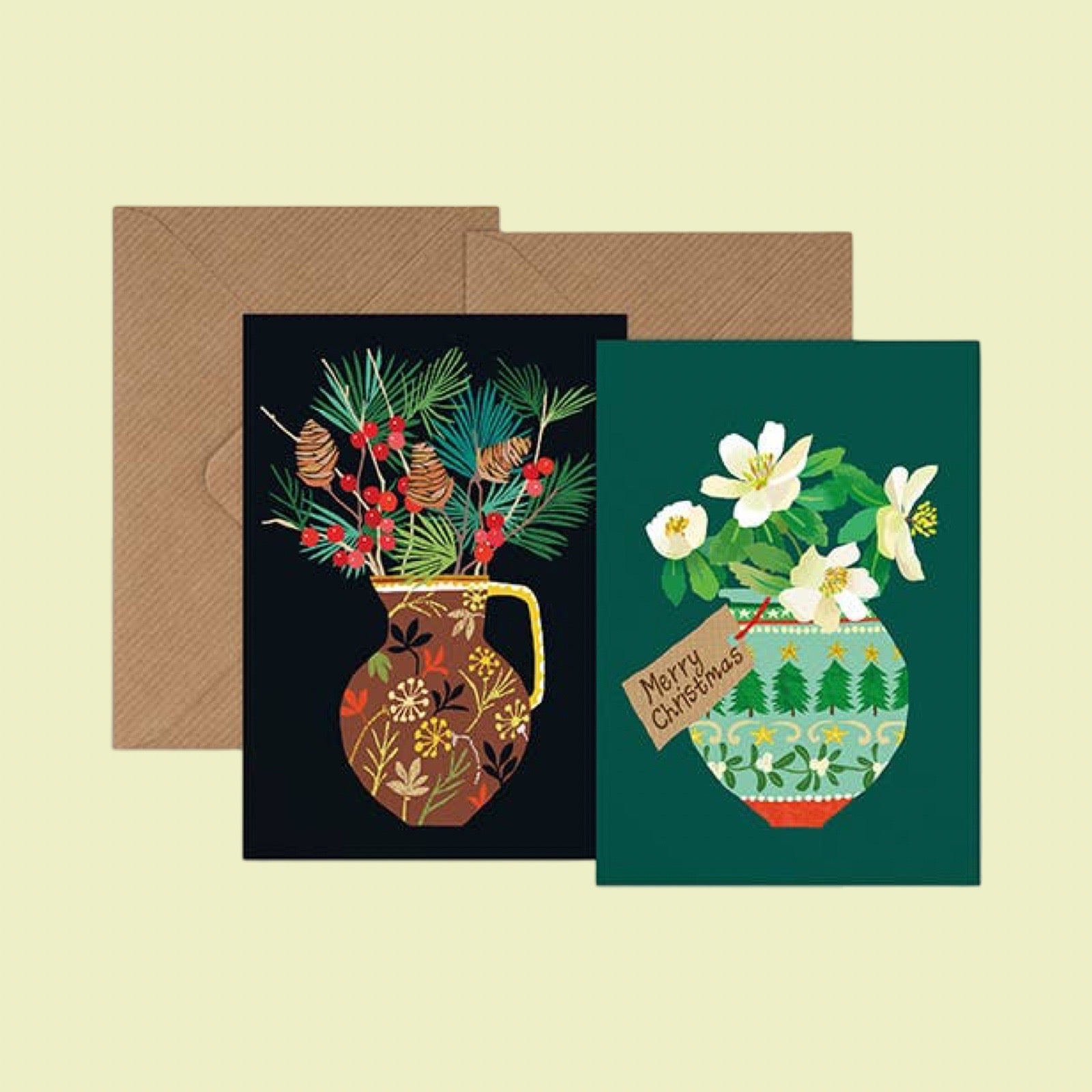Two Christmas greeting cards with illustrated floral vases and pine branches, shown with brown envelopes on a pale yellow background. One card has a black background with a brown vase and red berries, and the other has a dark green background with white flowers and a hanging 'Merry Christmas' tag.
