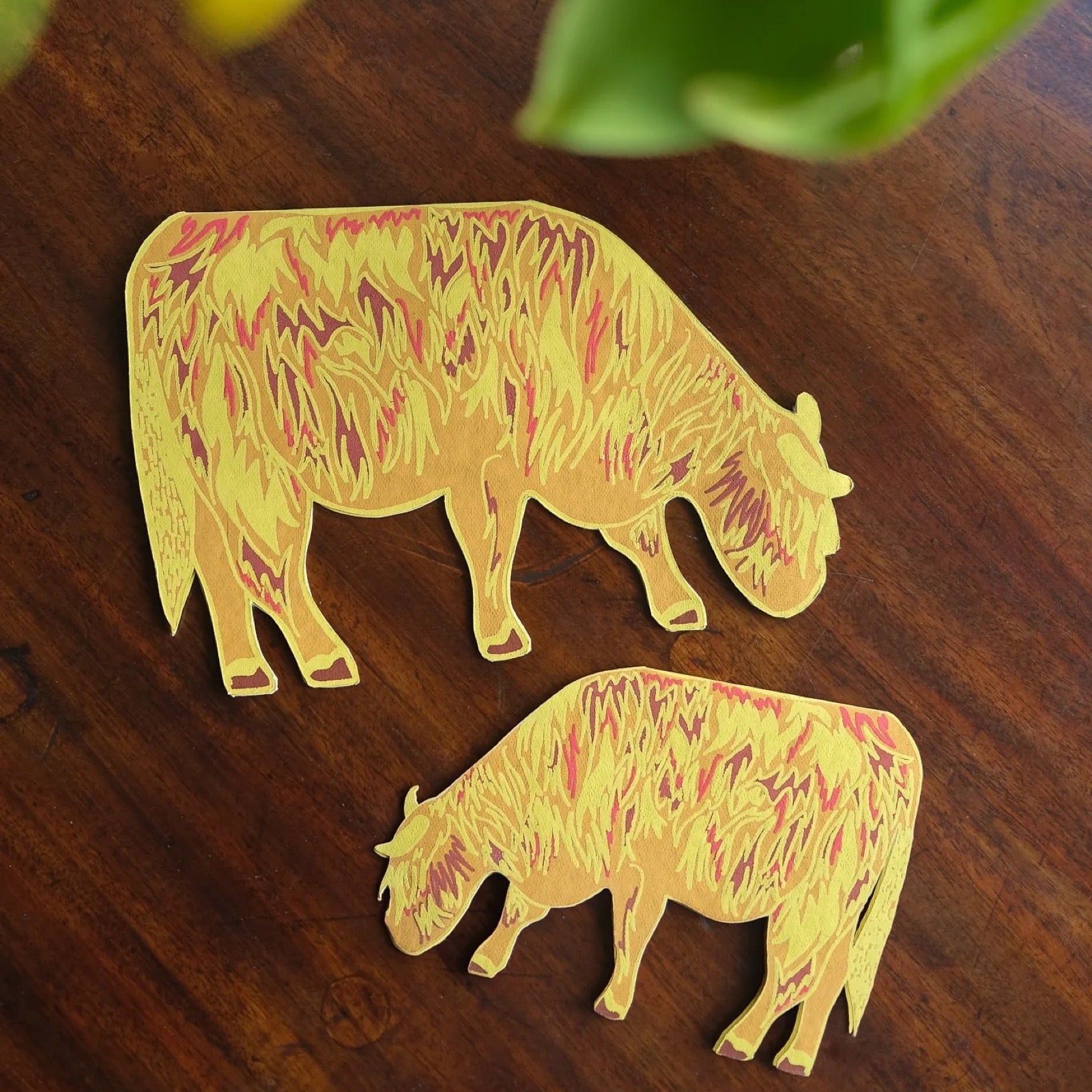 Two yellow highland cow shaped cards on a wooden surface with green leaves in the background.