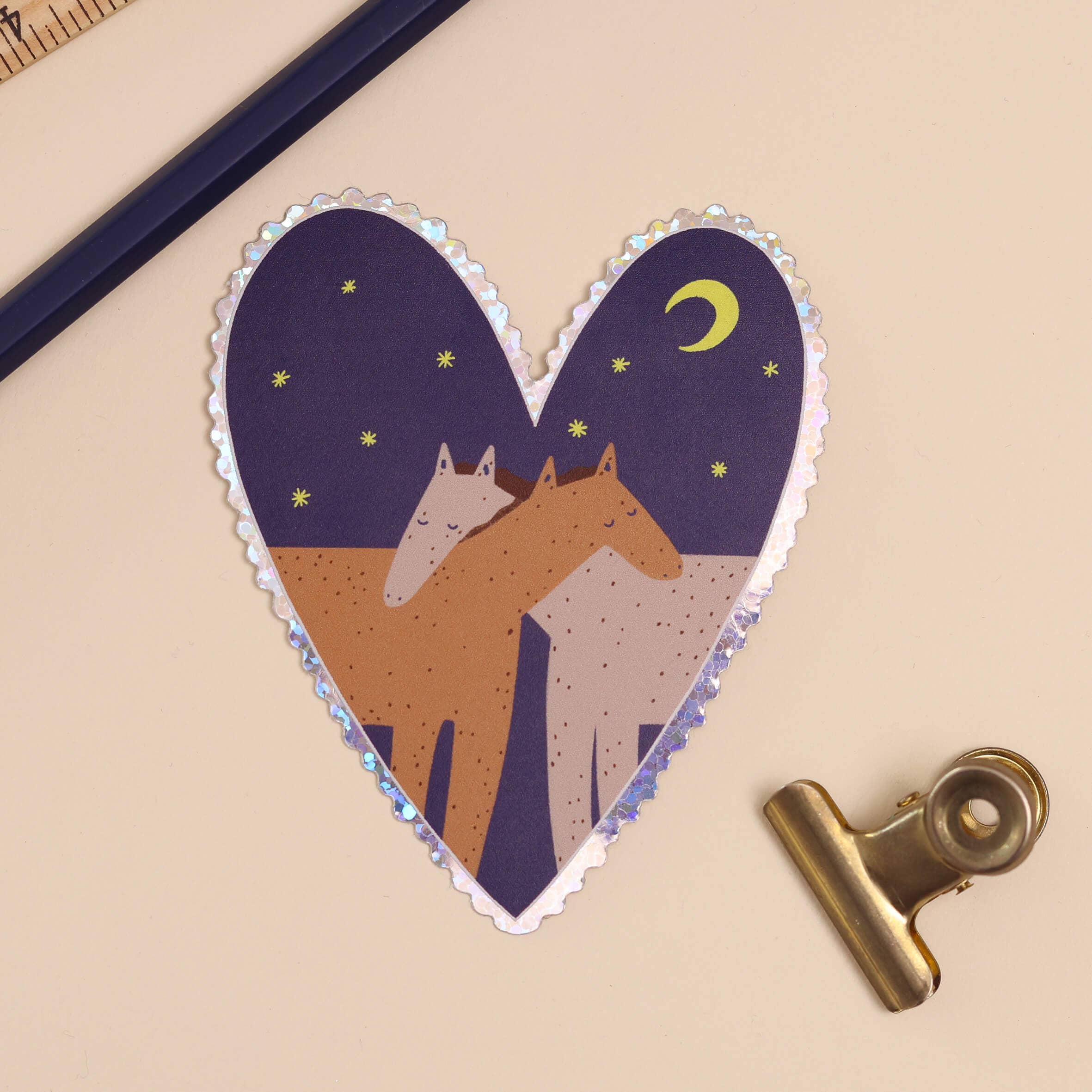 A heart-shaped sticker featuring an illustration of two horses under a crescent moon and stars, placed on a beige background with a gold binder clip and a dark blue pencil.