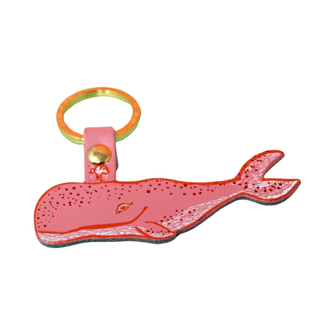 A humpback whale-shaped keyring in pink with a red outline, red embossed spots along the top and silver embossed dashes and the bottom and tail fin hanging from a gold-plated ring.
