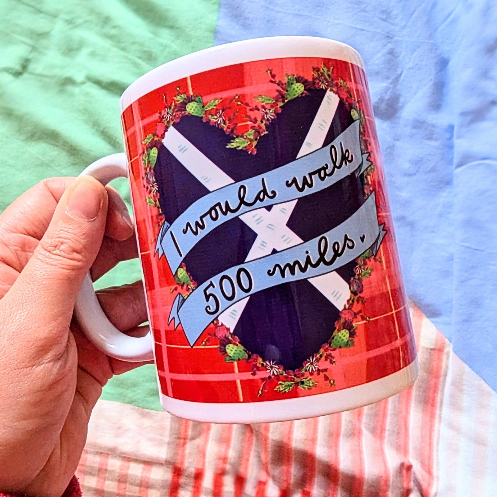 Hand holding a mug with a red tartan design and a Saltire heart with a banner with the text 'I would walk 500 miles' with a blurry fabric background.