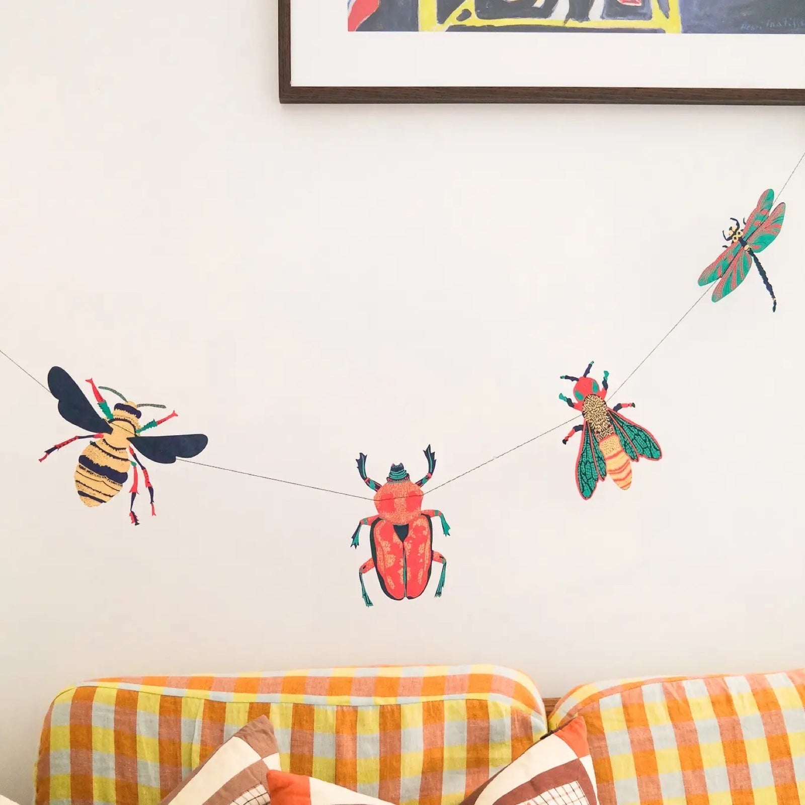 A paper garland with 10 screen printed insects, beetles, wasps, dragonflies and moths in shades of green, red and gold hanging against a white wall above a orange checked sofa.