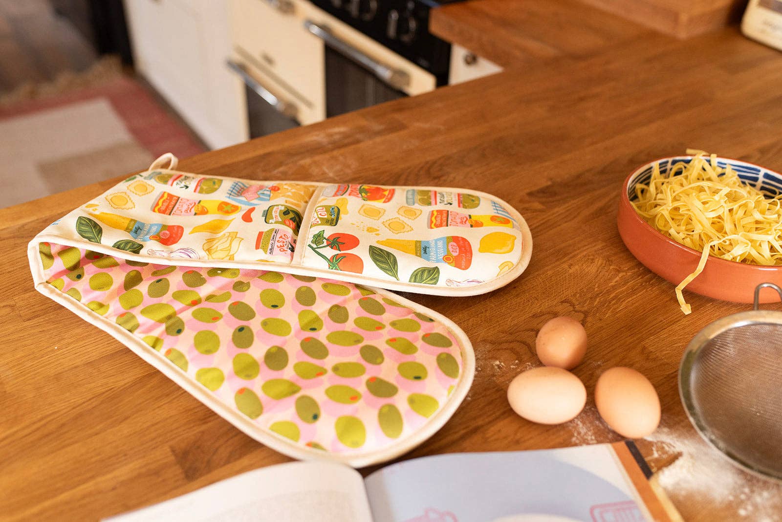 Colorful oven mitts and pot holders on a wooden kitchen counter with eggs and a bowl of pasta.