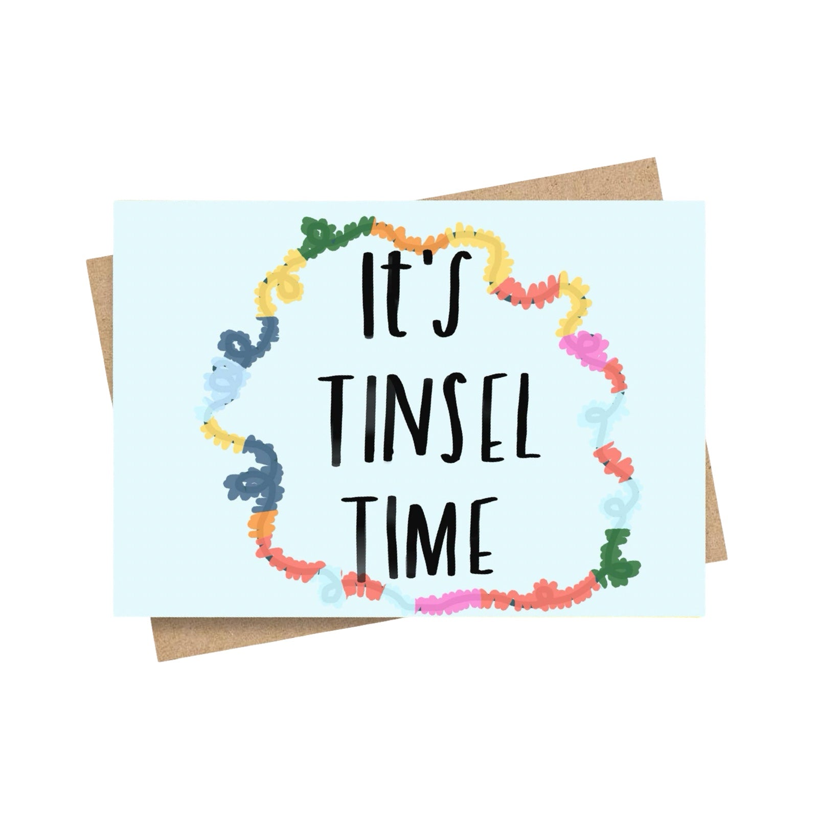 A greeting card with a light blue background and the phrase 'IT'S TINSEL TIME' in black handwritten-style text, surrounded by a colourful garland-like border. A brown envelope is partially visible behind the card on a white background.