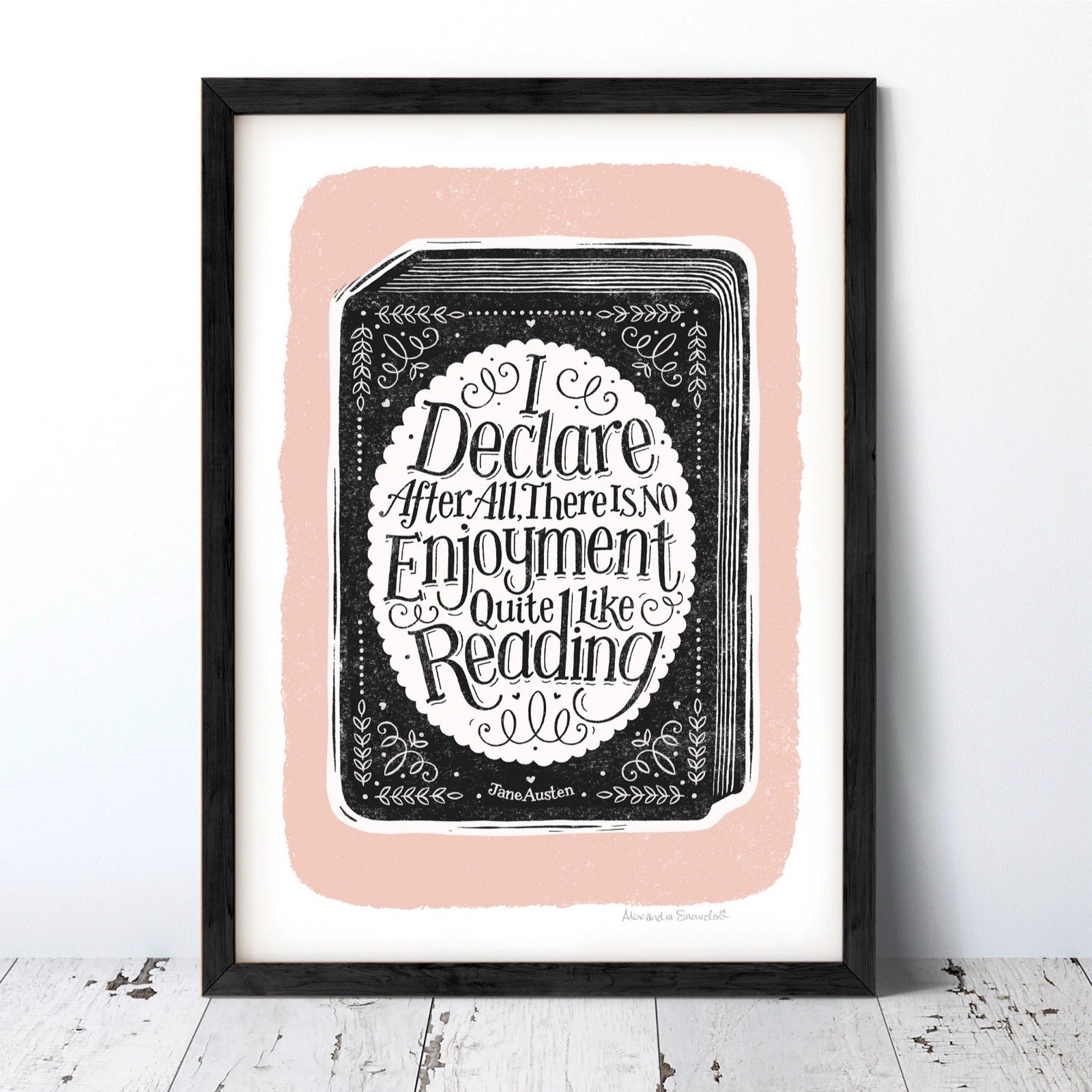 Framed print with decorative text on a book, on a pink background, leaning against a wall.