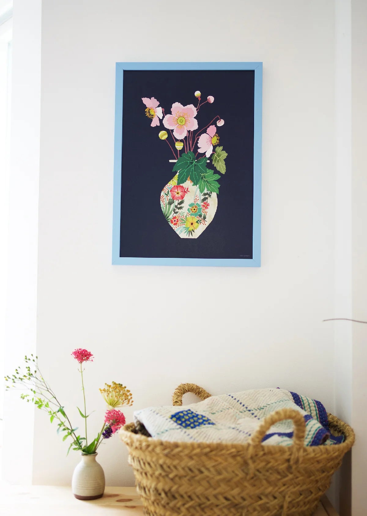 Framed floral artwork on a wall with a woven basket and flowers below.