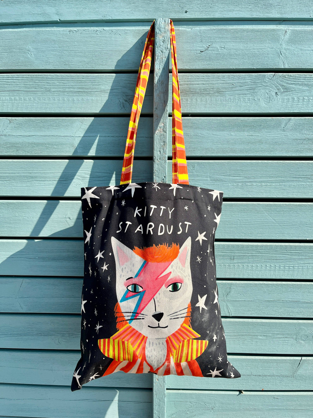 A black canvas tote bag featuring a cat illustration with lightning-bolt makeup and the text 'KITTY STARDUST,' hanging against a light blue wooden plank wall. The bag has orange and yellow striped handles.