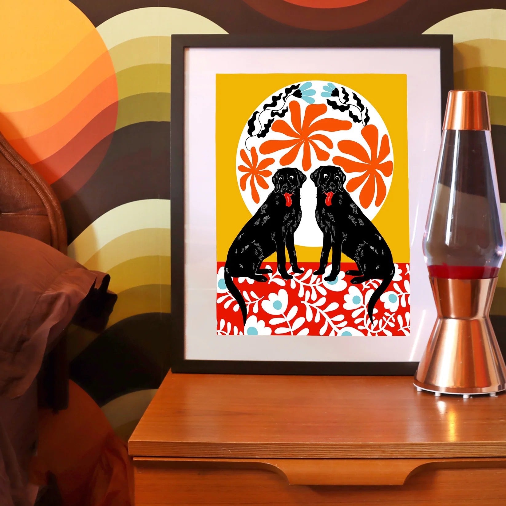 Labrador print featuring two black labs sitting on a red floral rug against a mustard background with a white patterned sun, on a bedside table next t a lava lamp and an abstract wall.