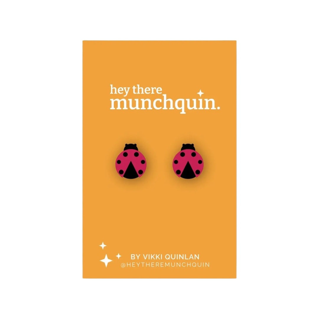 A pair of ladybird-shaped wooden stud earrings in red and black, with the brand name 'Munchquin' and the designer's name 'Viki Quinlan' printed on the image.
