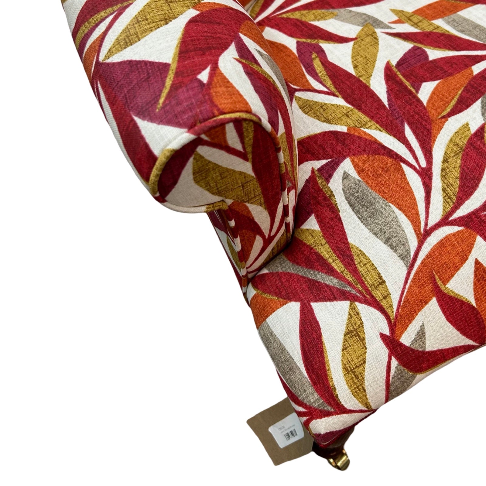 A hand-crafted armchair in cream linen with a leaves pattern in dark red, burnt orange, mustard and grey. The chair has a button-back and is finished with red binding, mahogany stained legs and gold castors.