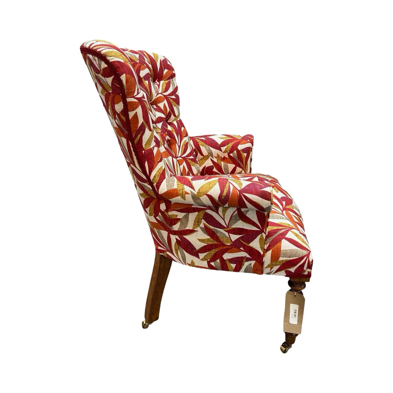 A hand-crafted armchair in cream linen with a leaves pattern in dark red, burnt orange, mustard and grey. The chair has a button-back and is finished with red binding, mahogany stained legs and gold castors.