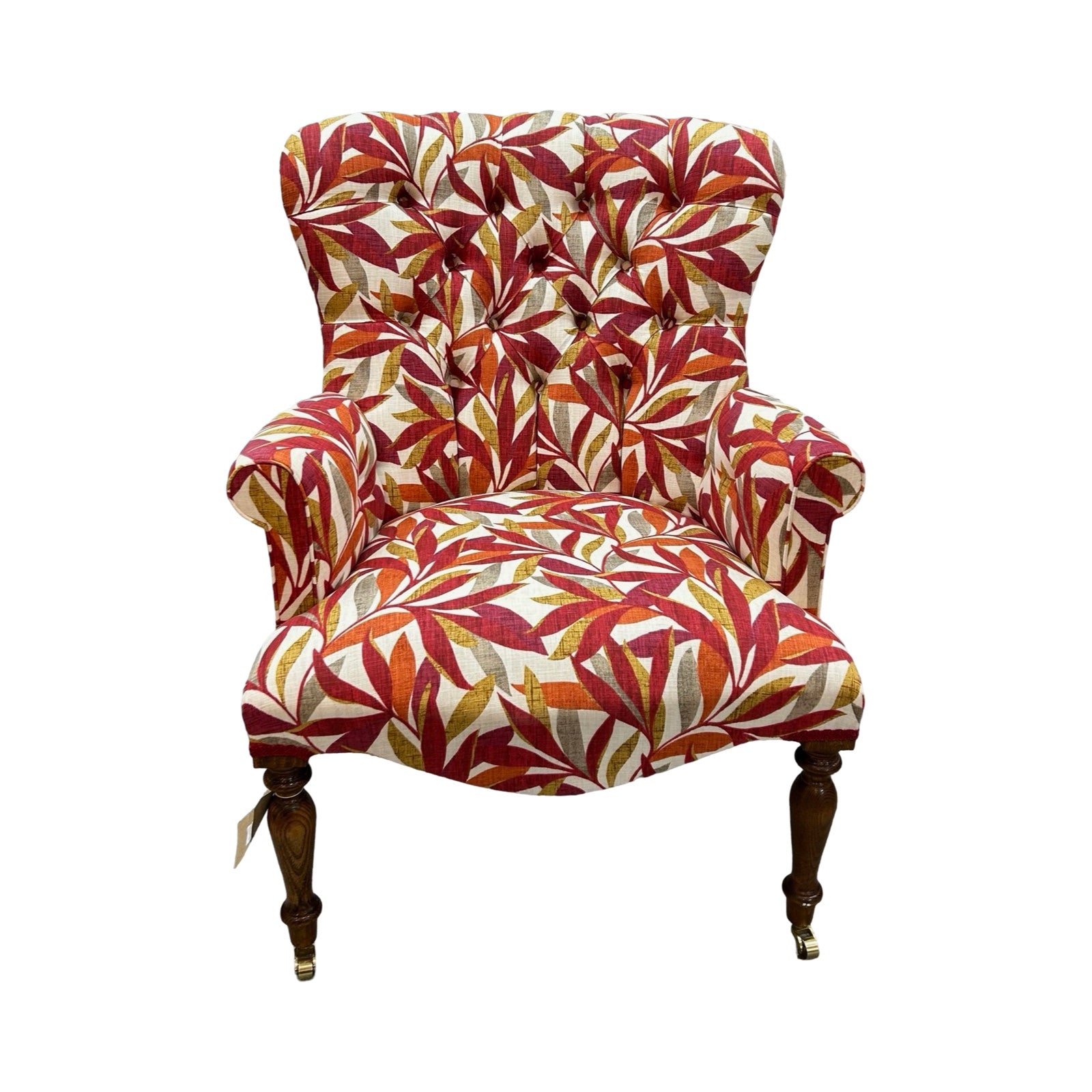 A hand-crafted armchair in cream linen with a leaves pattern in dark red, burnt orange, mustard and grey. The chair has a button-back and is finished with red binding, mahogany stained legs and gold castors.