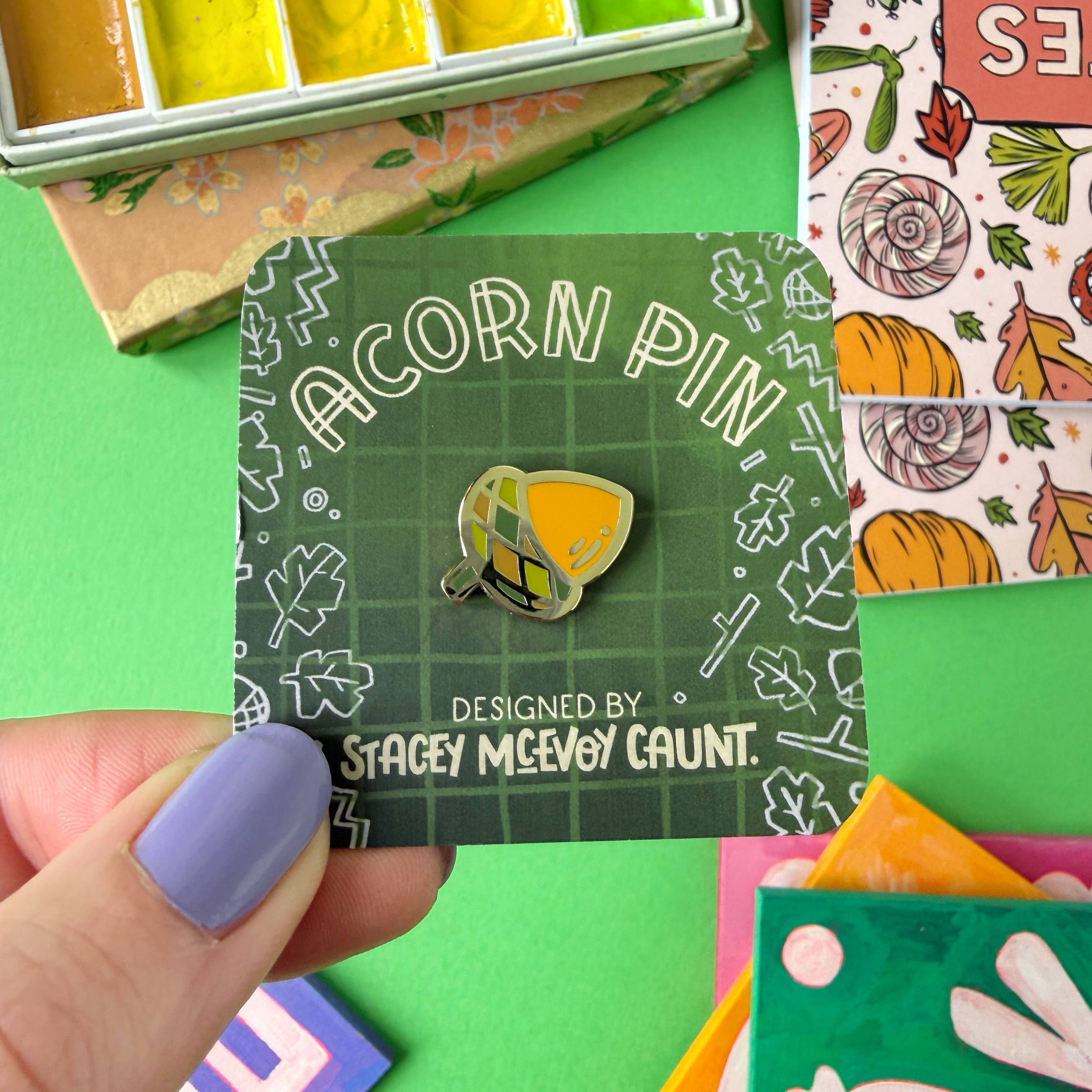 Acorn pin on a card with 'Stacey McEvoy Caunt' design, surrounded by colourful items on a green background.