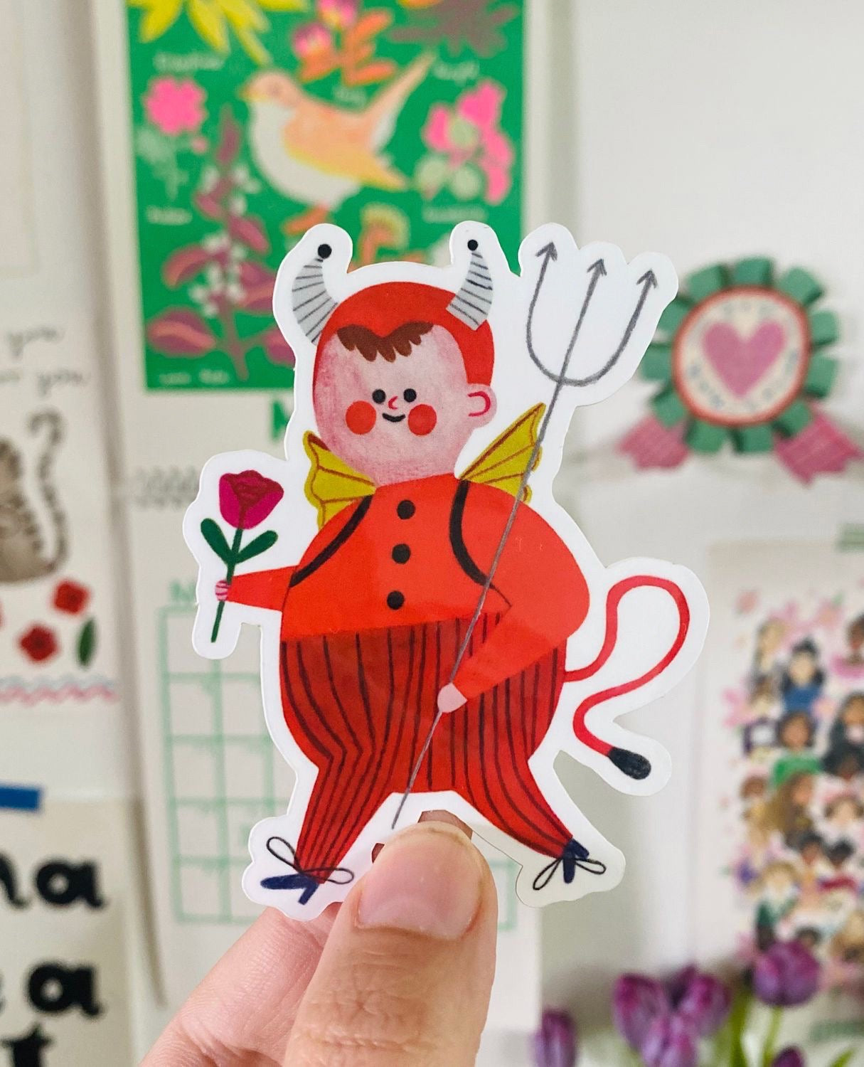 Sticker of a cartoonish devil character with horns and a pitchfork held by a hand, with a colourful background.