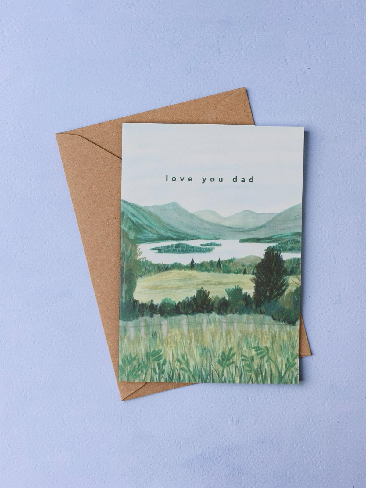 Greeting card with landscape illustration and 'love you dad' text, placed with a brown envelope against a light blue background.