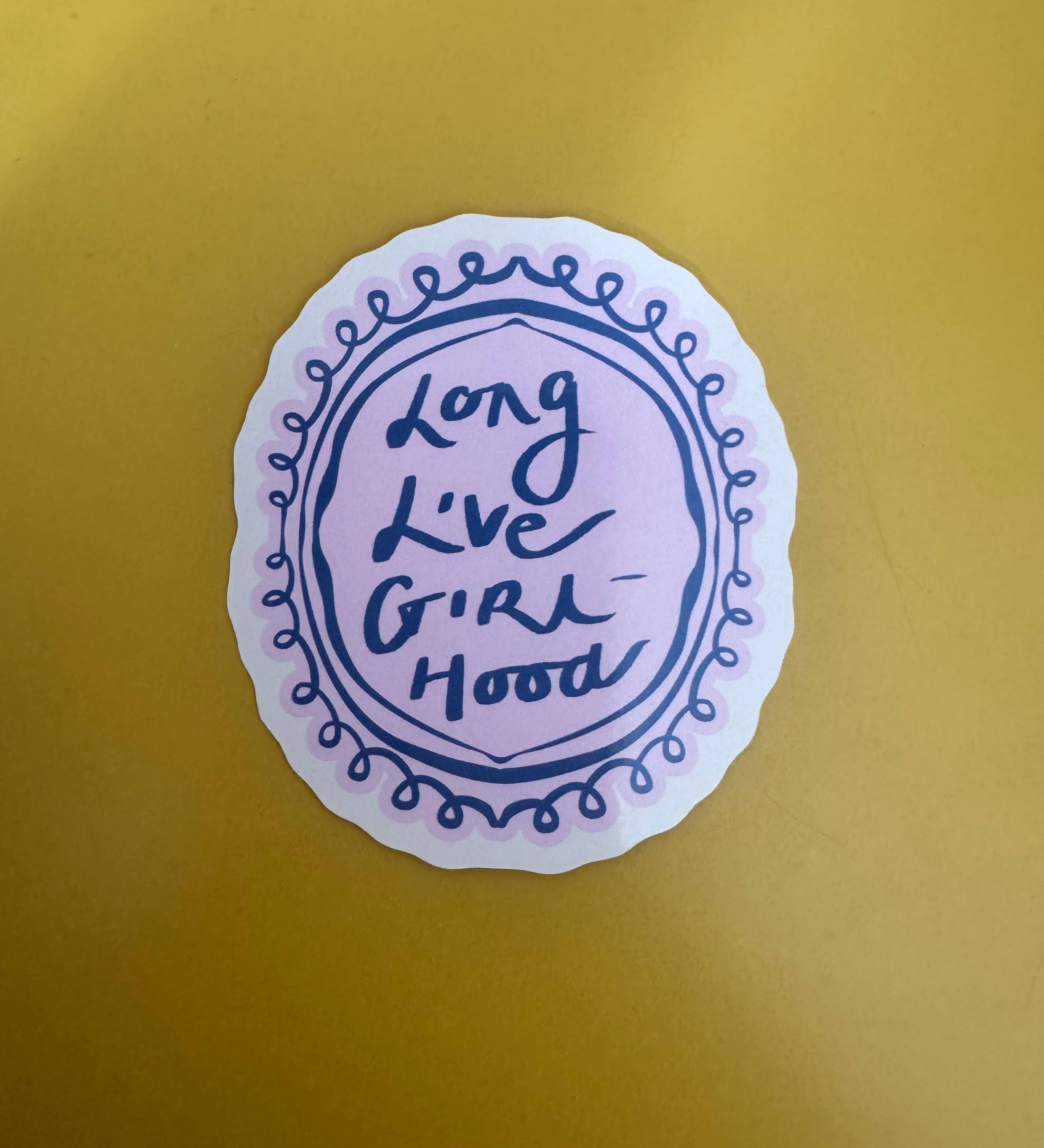 A round sticker with a scalloped white border and a lavender-pink center, featuring the handwritten phrase 'long live girl hood' in dark blue ink, placed on a solid mustard-yellow background.