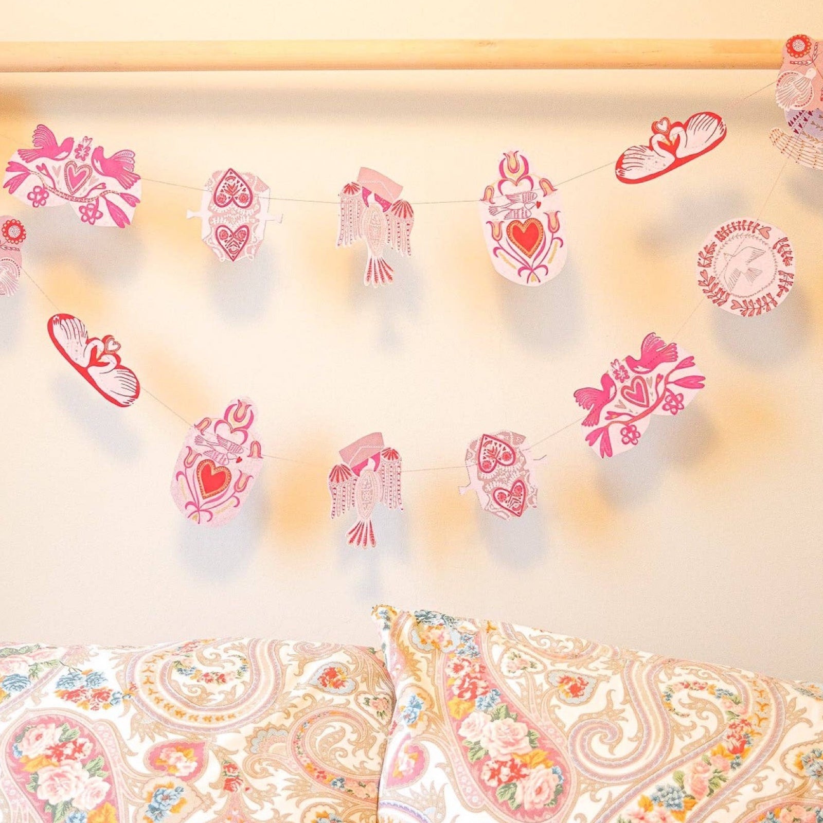 A paper garland with sixteen screen-printed folk bird designs in shades of pink, red and white hanging on a wooden bed frame.