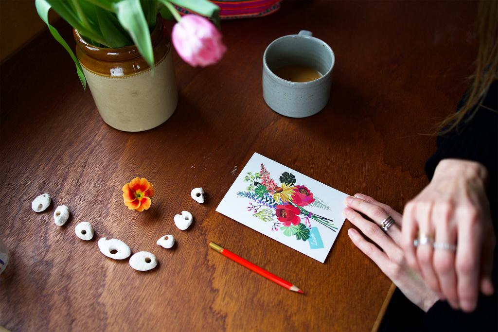 Person holding a card with floral design on a wooden table with flowers and a mug.