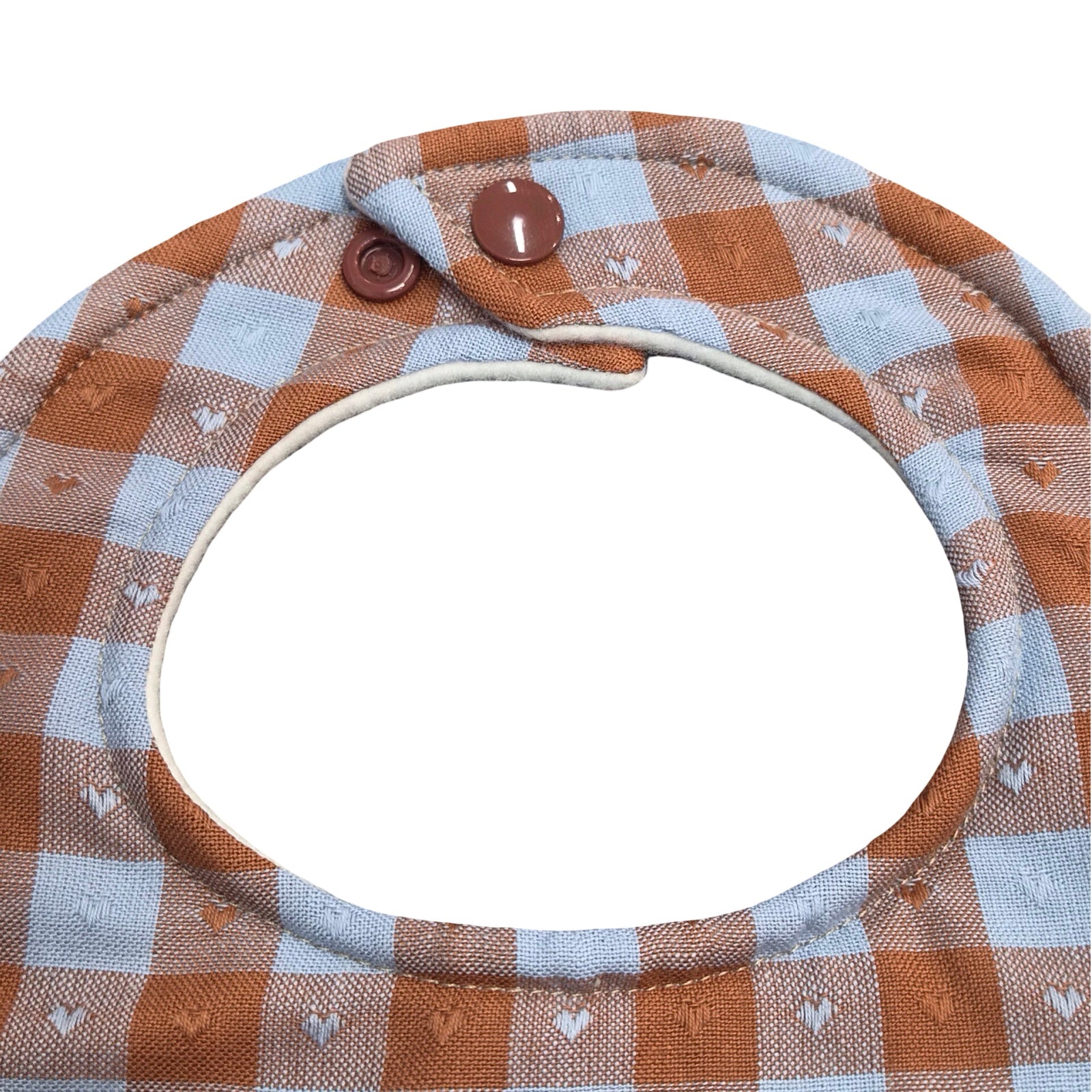 Close-up of a baby bib with a brown and light blue gingham pattern, featuring small embroidered hearts and white snap buttons at the top.
