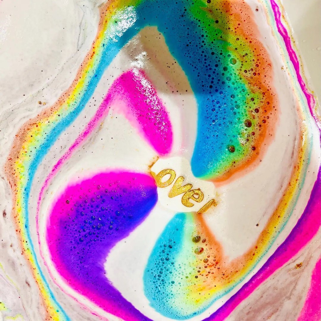 A white love heart shaped bath bomb with the text 'Love Is' written in the centre in gold dissolving in the bath leaving a rainbow of colours