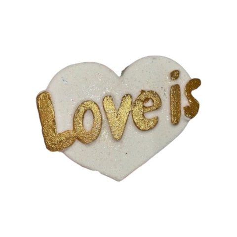 A white love heart shaped bath bomb with the text 'Love Is' written in the centre in gold.