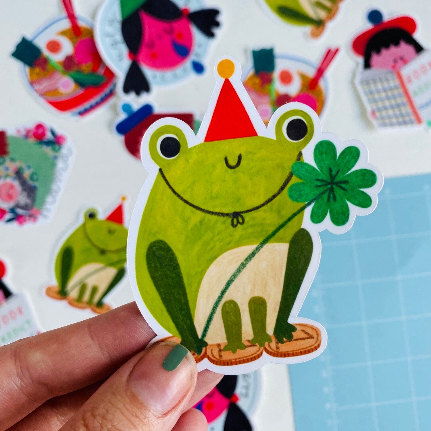 Hand holding a green frog sticker with a party hat and flower, surrounded by other colourful stickers.