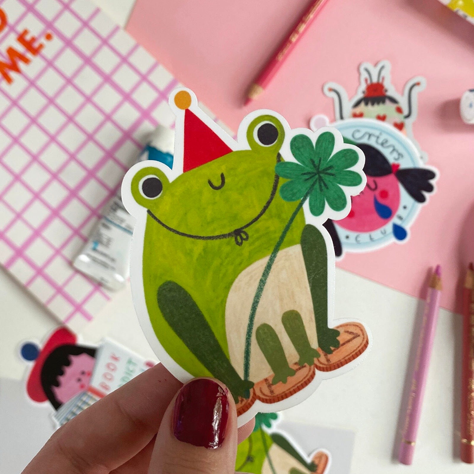 Hand holding a sticker of a green frog with a four-leaf clover on a pink background with stationery items.