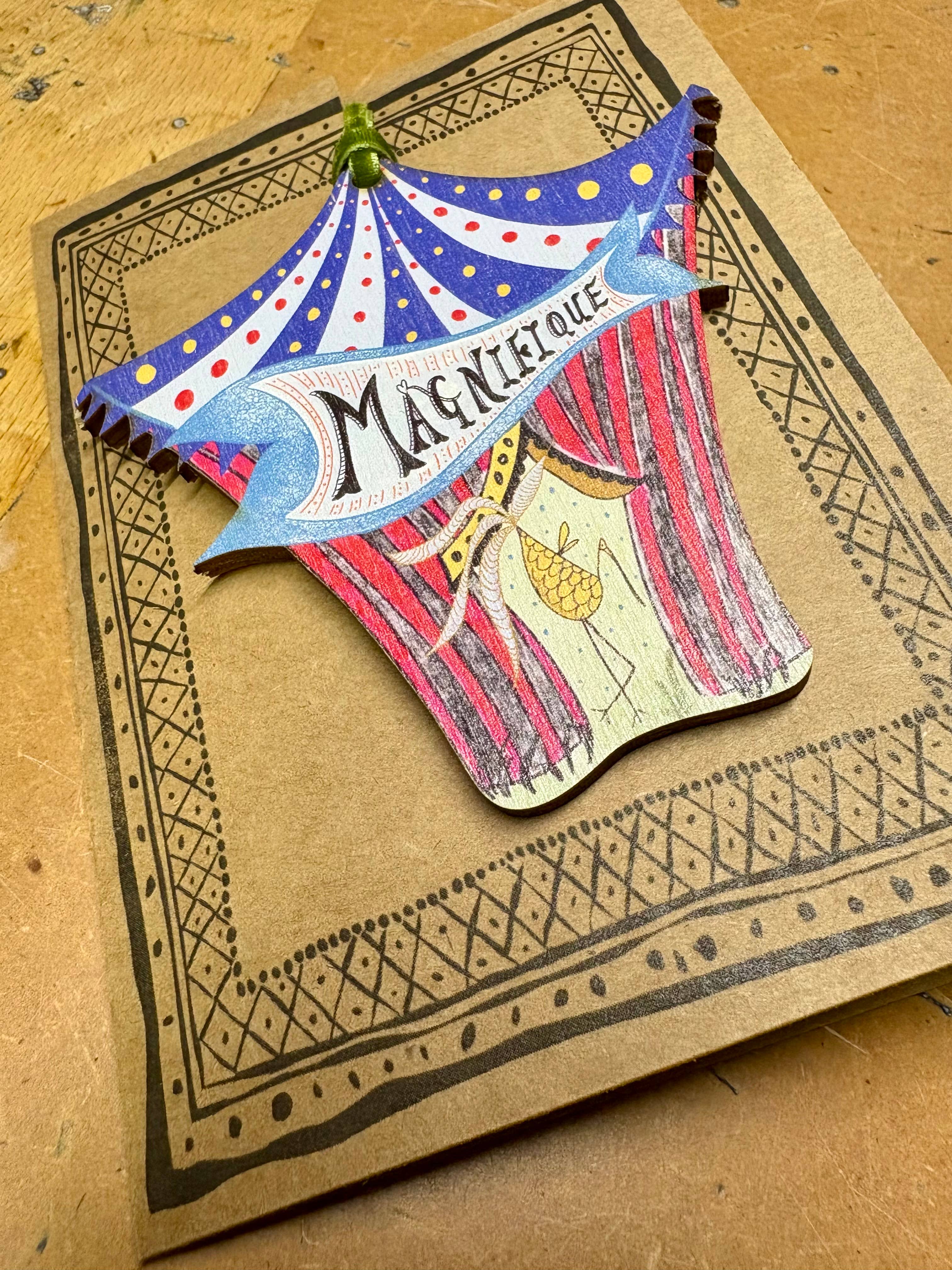 A handcrafted wooden decoration shaped like a vintage circus tent, featuring painted red and blue stripes, polka dots, curtains, and a banner reading 'MAGNIFIQUE', displayed on a patterned backing card on a wooden surface.