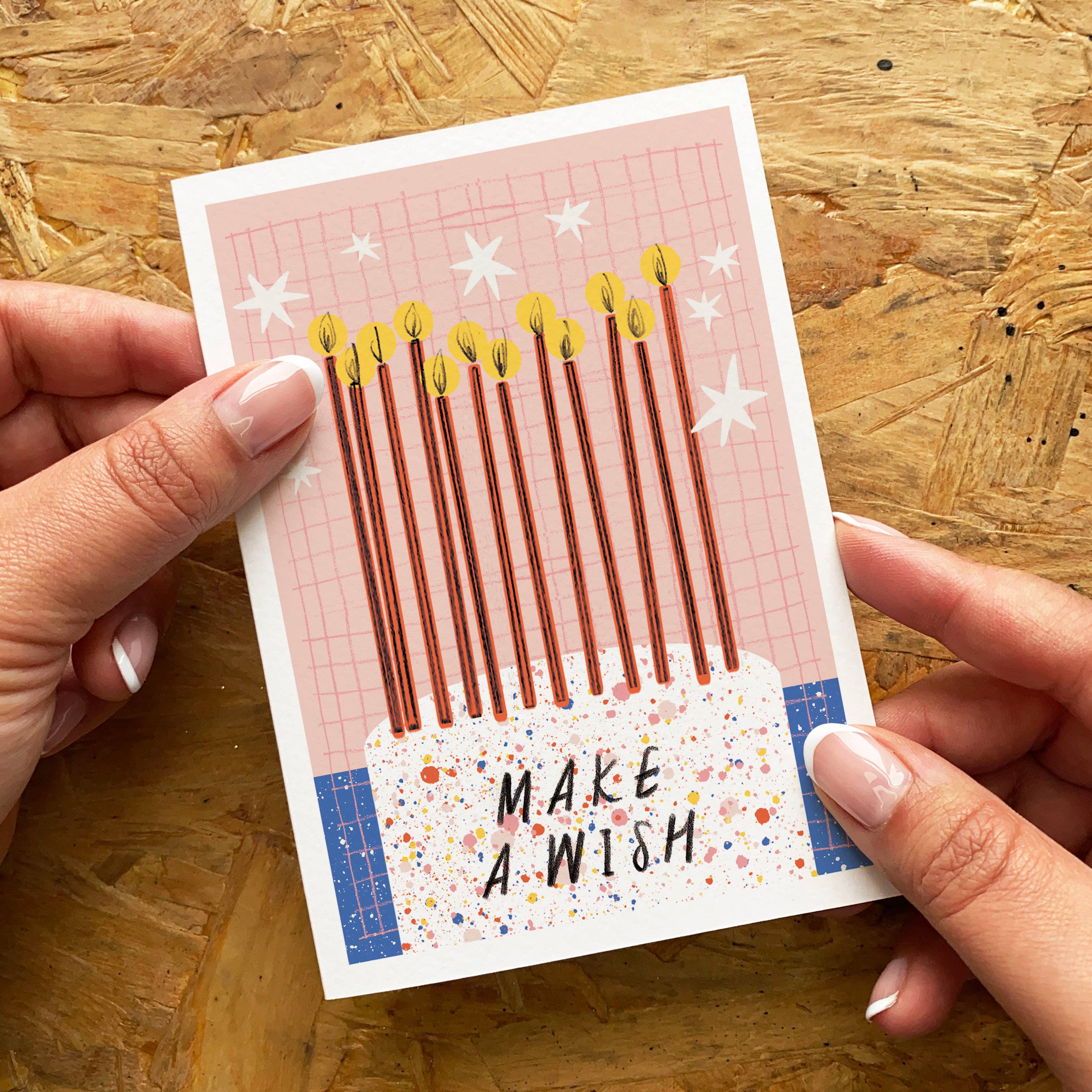 Card with candles and 'Make a Wish' text held by hands on a wooden surface.