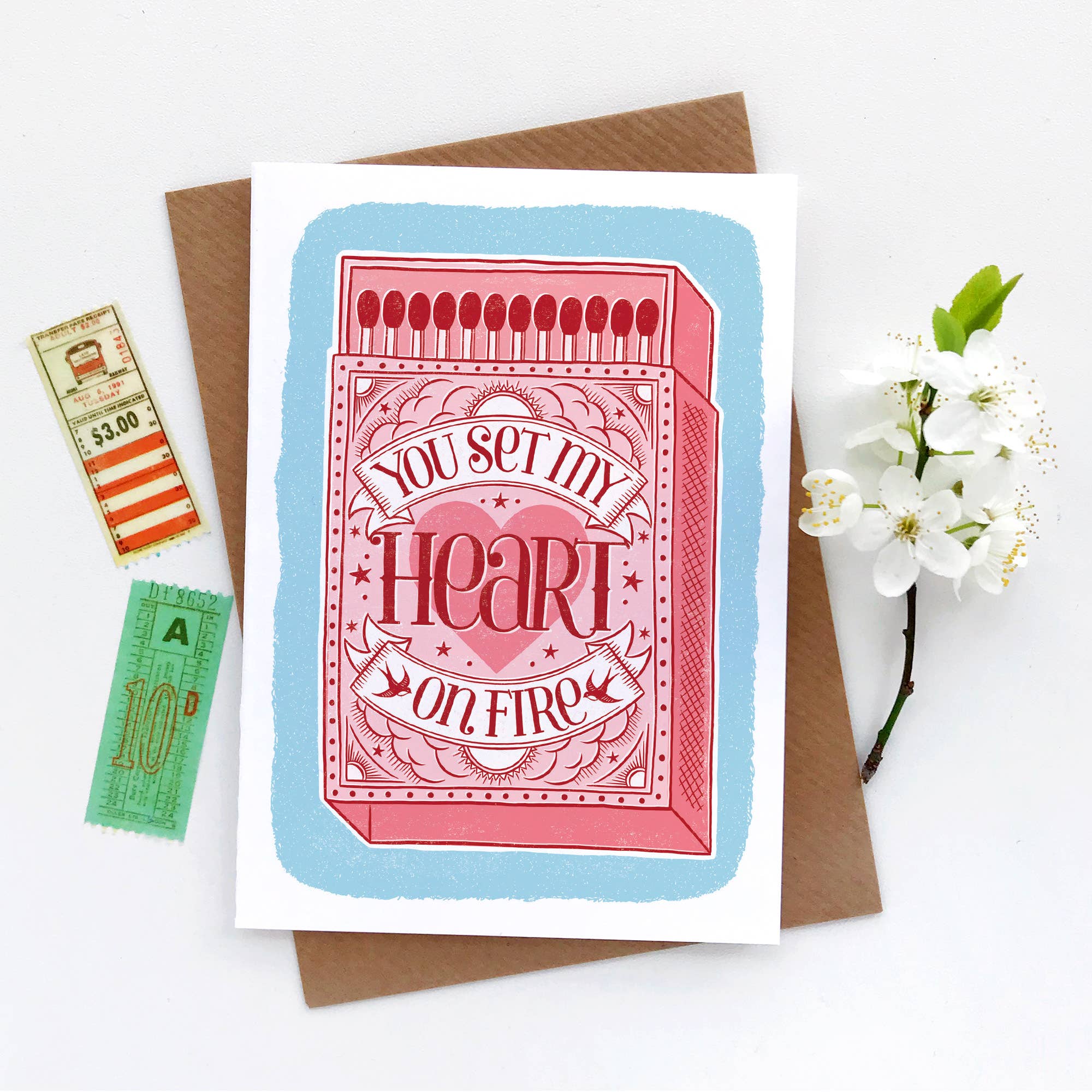Greeting card with matchbox design and romantic text, placed on a white surface with a brown envelope and decorative items.