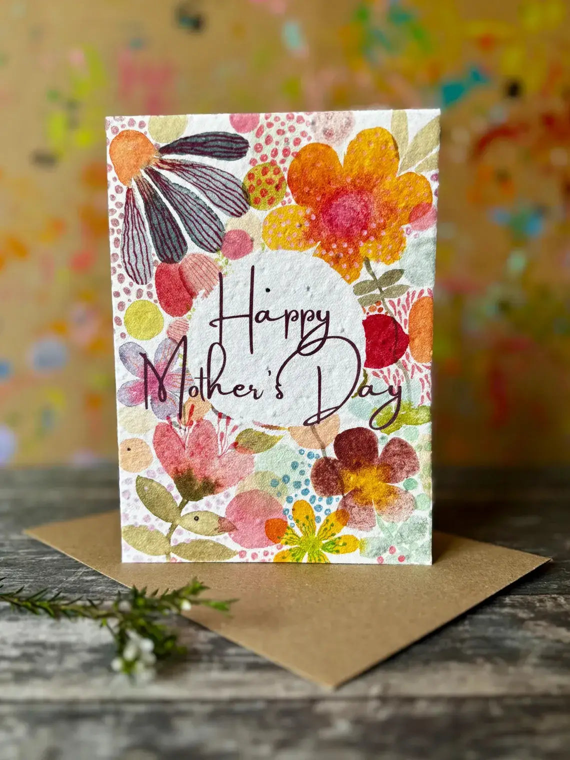 A Mother's Day greeting card with a colourful watercolour floral design and the words 'Happy Mother's Day' in black script, standing upright on a brown envelope over a rustic wooden surface.