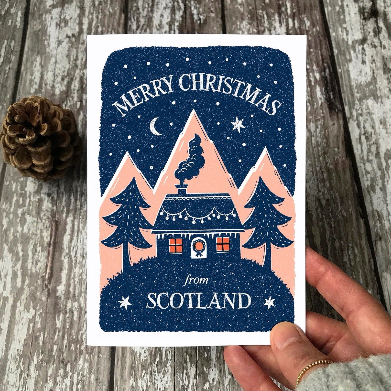 Christmas card with a house and trees design, held by a hand on a wooden surface.
