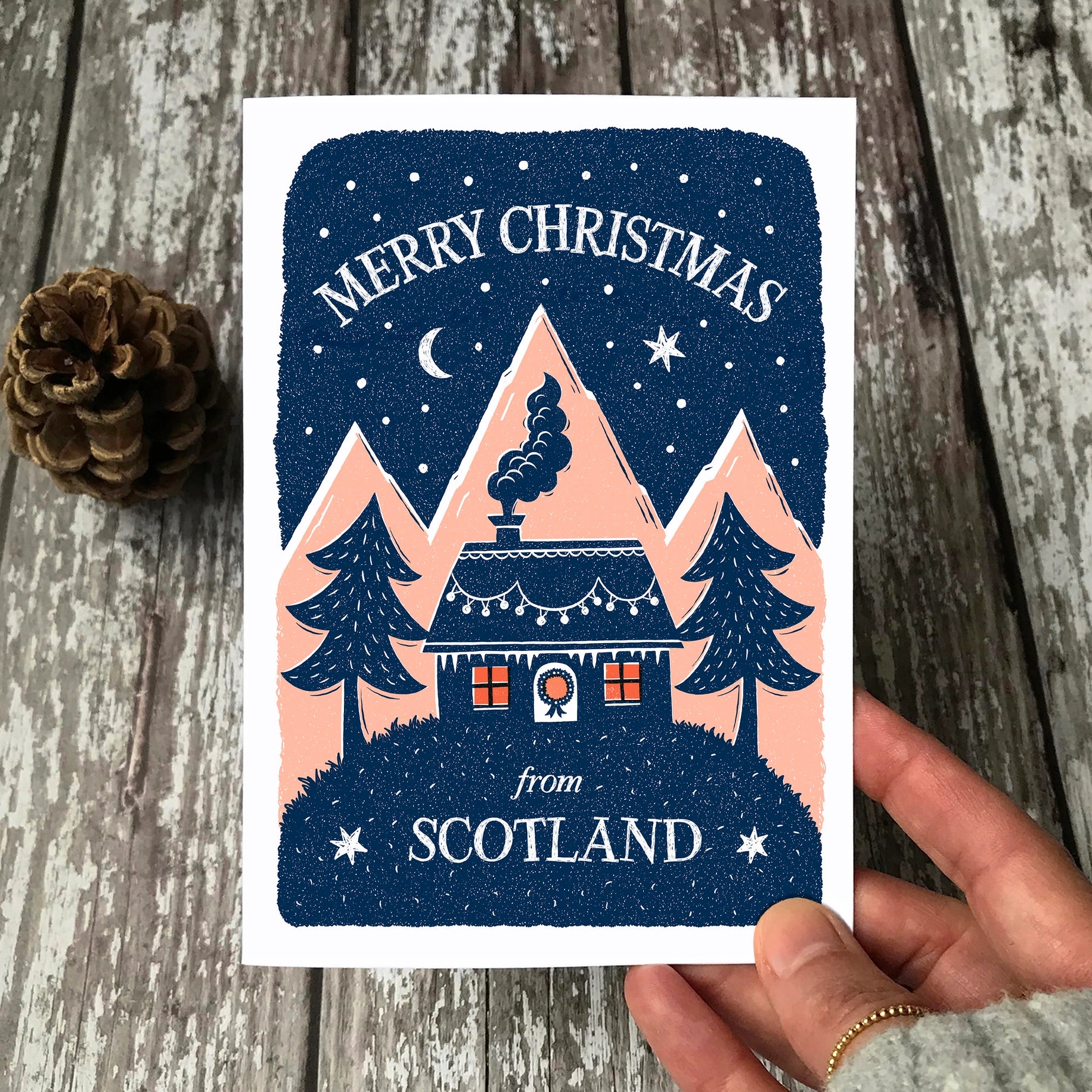 Christmas card with a house and trees design, held by a hand on a wooden surface.