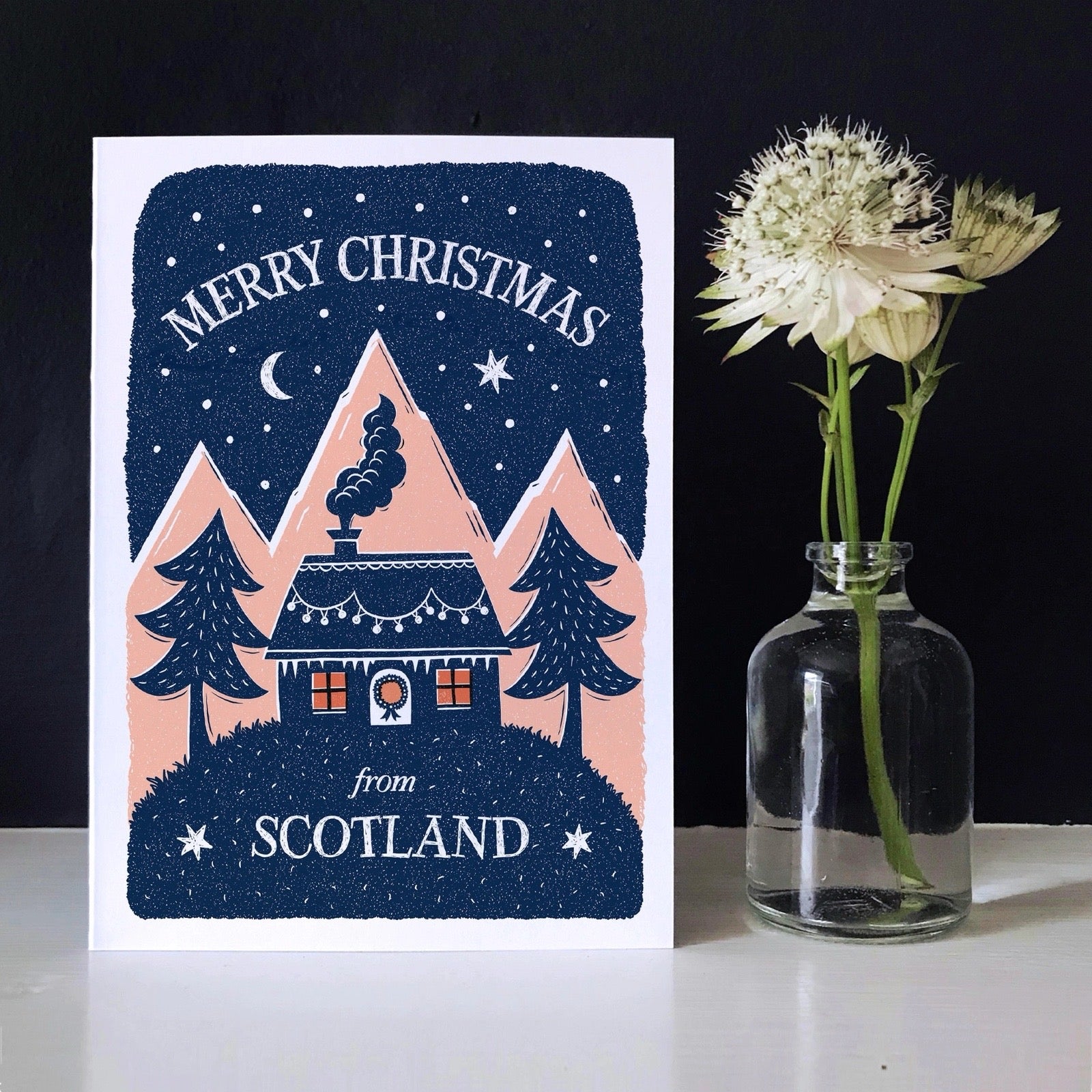 Christmas card with a house and trees design, next to a vase with flowers on a dark background.