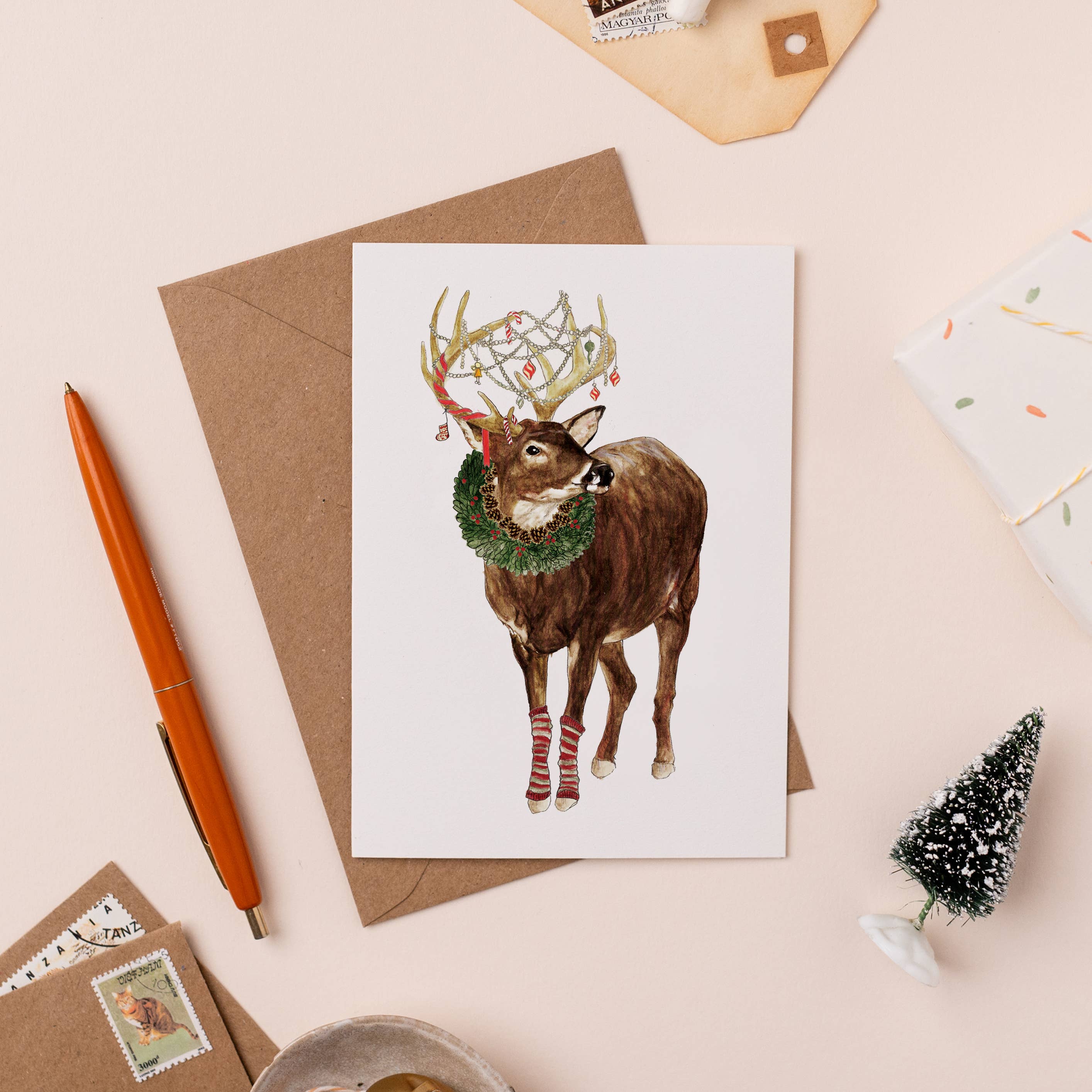 Greeting card with a reindeer illustration on a beige surface with stationery items.