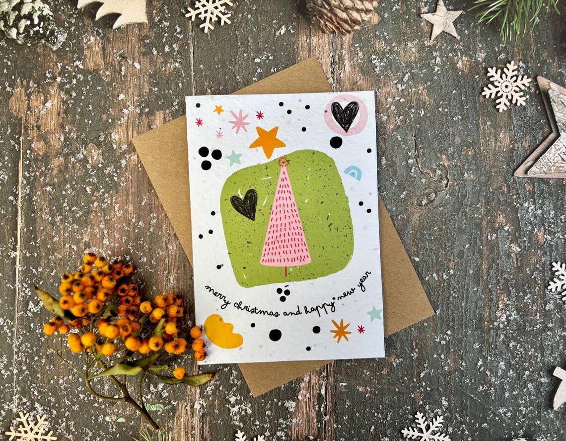 A greeting card with a whimsical Christmas tree illustration, placed on top of a brown kraft envelope over a rustic wooden surface with decorative snowflakes and orange berries.