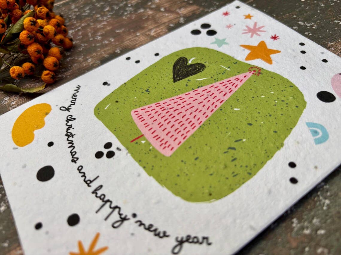 A handmade paper greeting card with a whimsical New Year illustration, featuring a pink tree on a green background with the text 'merry christmas and happy new year'. The card is displayed on a rustic wooden surface next to dried orange berries.