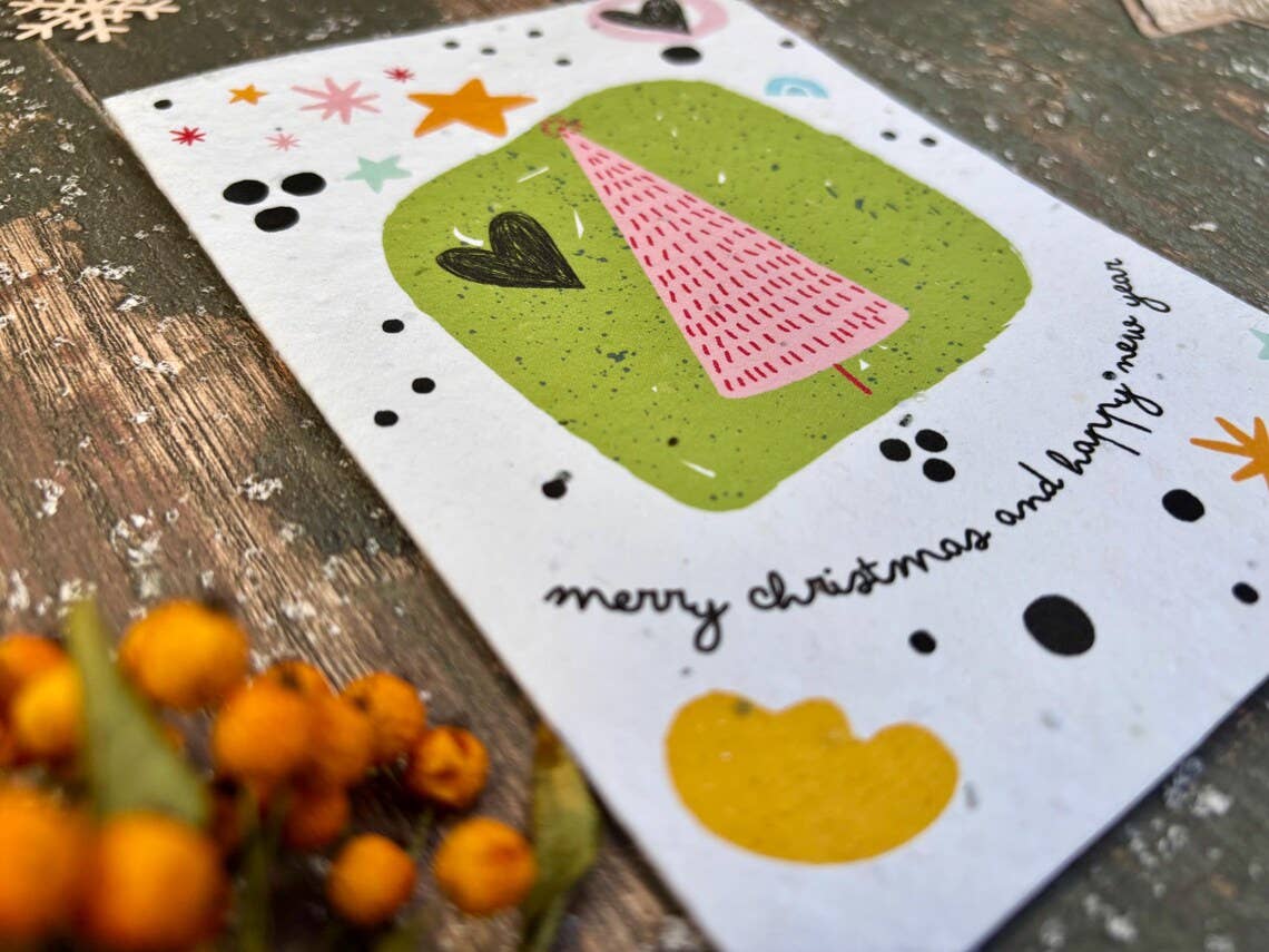 A handmade Christmas greeting card with a green tree illustration and the text 'merry christmas and happy new year', styled on a rustic wooden surface with small orange berries in the foreground.